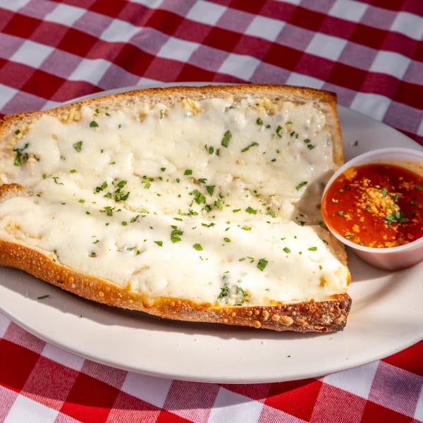 Cheesy garlic bread with parsley and a side of marinara sauce on a white plate and red checkered tablecloth.