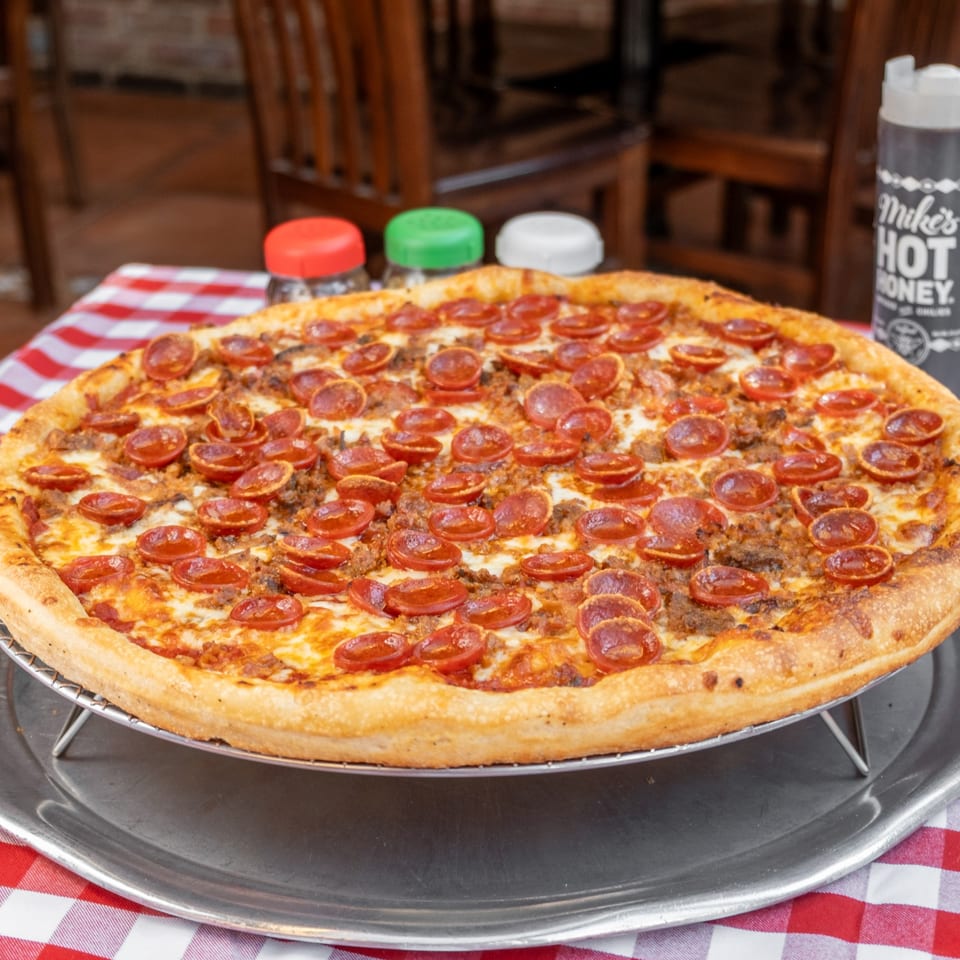 A pepperoni and sausage pizza on a metal stand, with hot honey and shakers in the background.