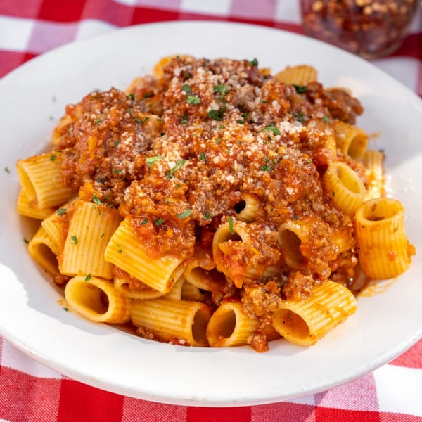 Rigatoni pasta with meat sauce, parmesan cheese, and parsley on a white plate over a red and white checkered tablecloth.